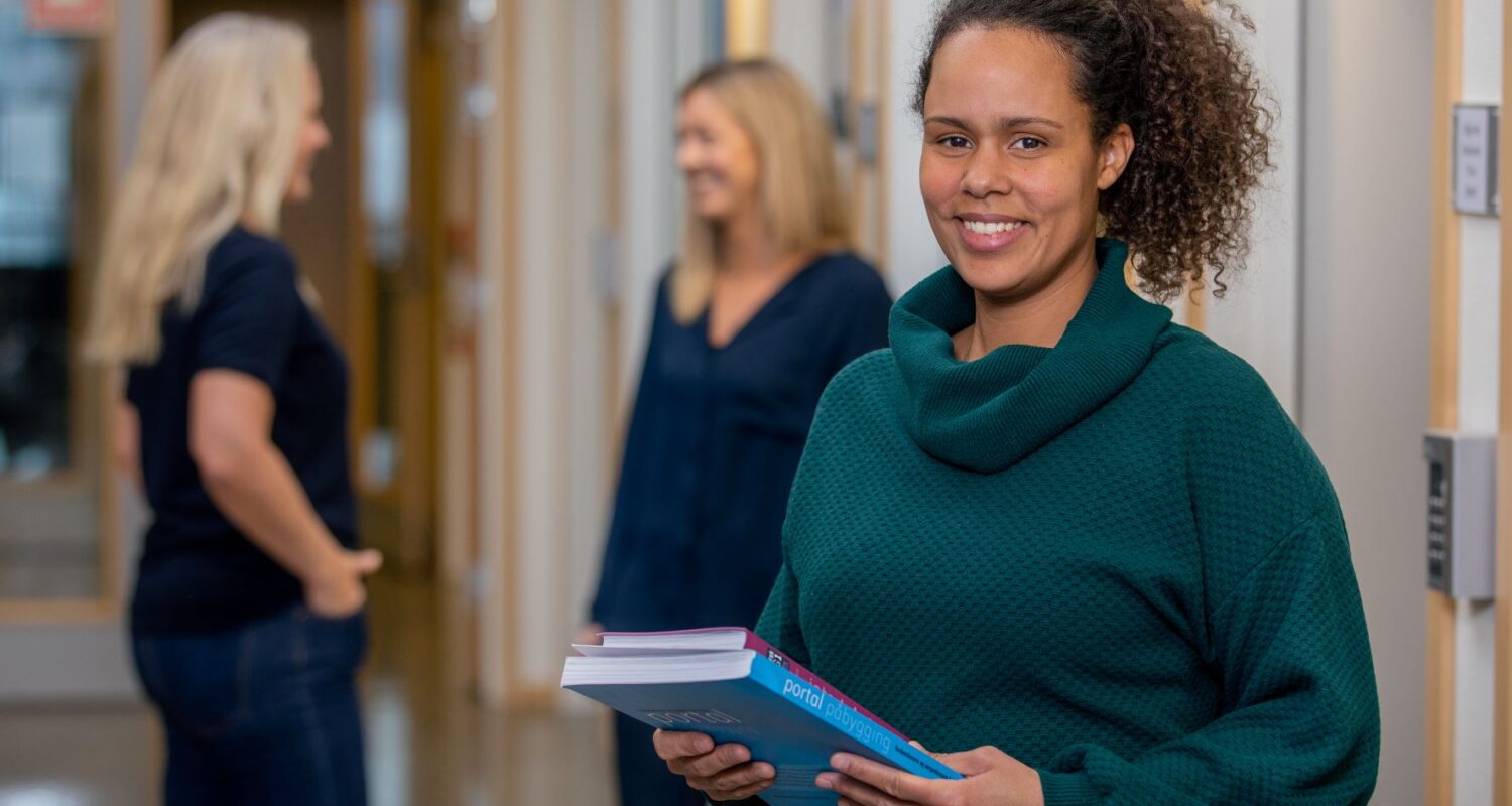 Admaya-Feiring_2 En kvinne med bøker i hånden står i en gang og smiler til kameraet. To andre kvinner snakker sammen i bakgrunnen.