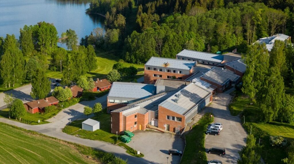 Feiring-4-1024x571 Flyfoto av et stort bygningskompleks i murstein med flere hustak, omgitt av trær, gressplener, parkerte biler og en innsjø i nærheten.