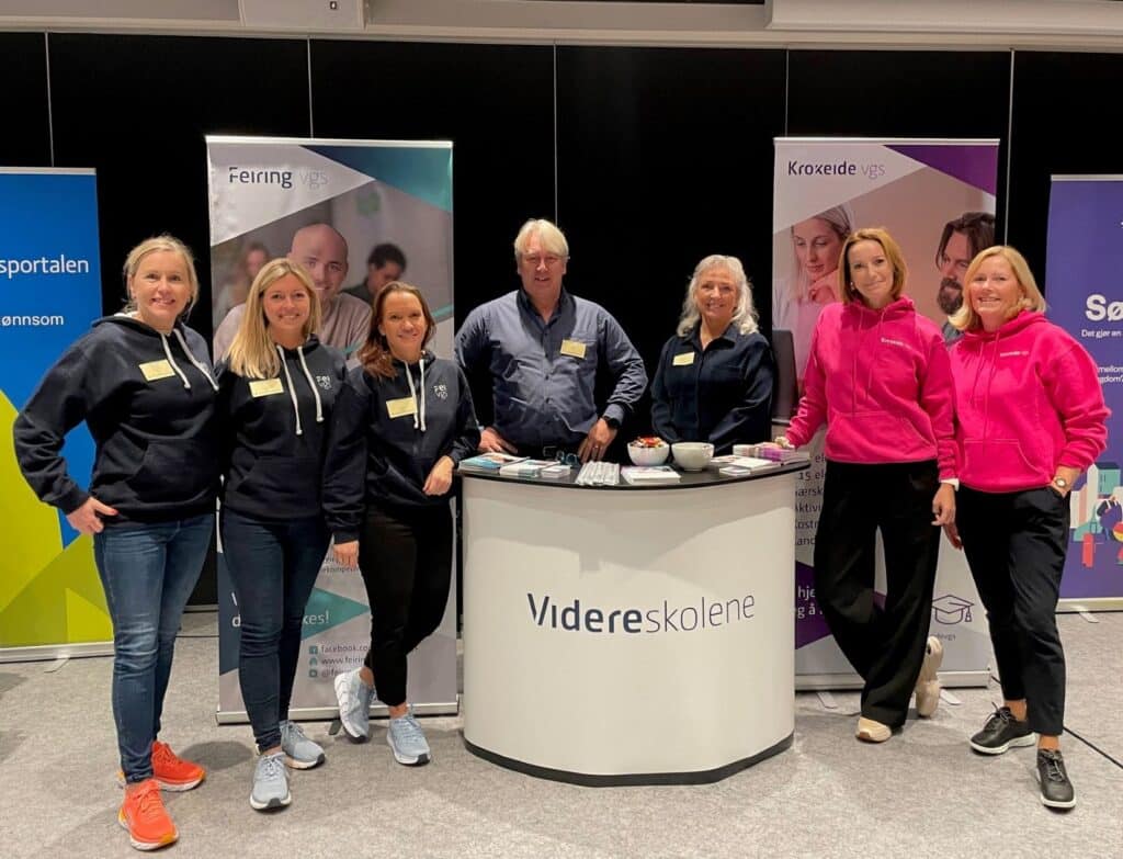 Folkehelsekonferansen-2022-3 Sju personer står bak et buet bord med "Videreskolene" på forsiden og poserer for et gruppebilde på et innendørsarrangement eller en messe.