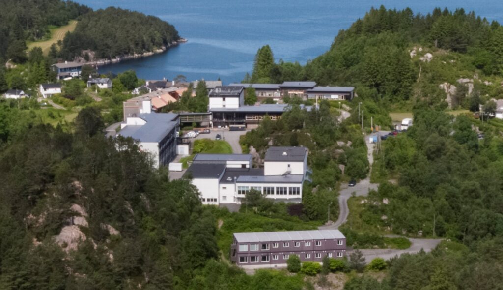 Krokeide-vgs-2-1024x590 Flyfoto av en gruppe bygninger omgitt av tette, grønne trær, med et vann og skogkledde åser i bakgrunnen.