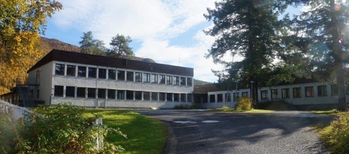 En rektangulær bygning i to etasjer med mange vinduer er omgitt av trær og grøntområder under en delvis overskyet himmel.