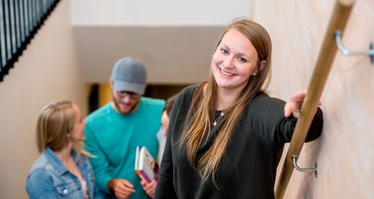 Sigri En ung kvinne står på et trapperekkverk og smiler til kameraet, mens tre personer i bakgrunnen snakker og holder bøker.