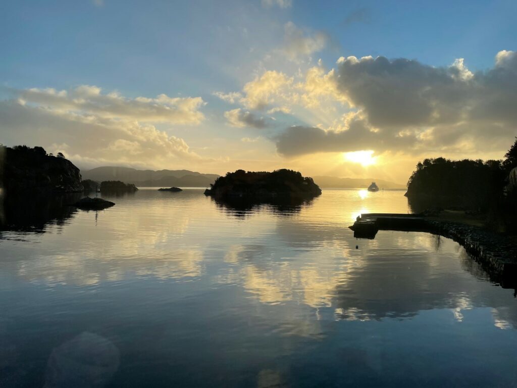 Utsikt-fra-Lyngvik-1-1024x768 En rolig vannflate speiler himmelen og skyene i solnedgangen, med små øyer og fjerne fjell i bakgrunnen.