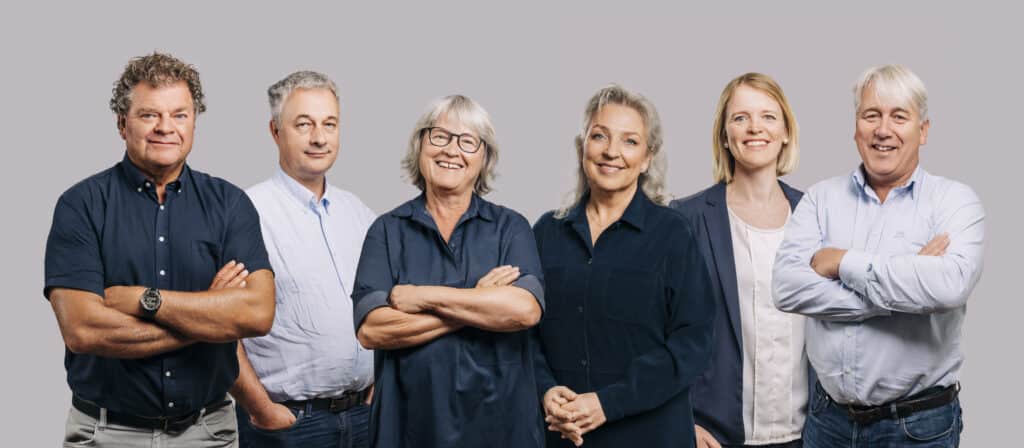 Videre-skolene_gruppebilde_WEB_copyright-Pernille-Sommer-1 Seks voksne står på rekke og rad mot en ensfarget bakgrunn, alle er kledd i blå eller marineblå klær og smiler til kameraet.