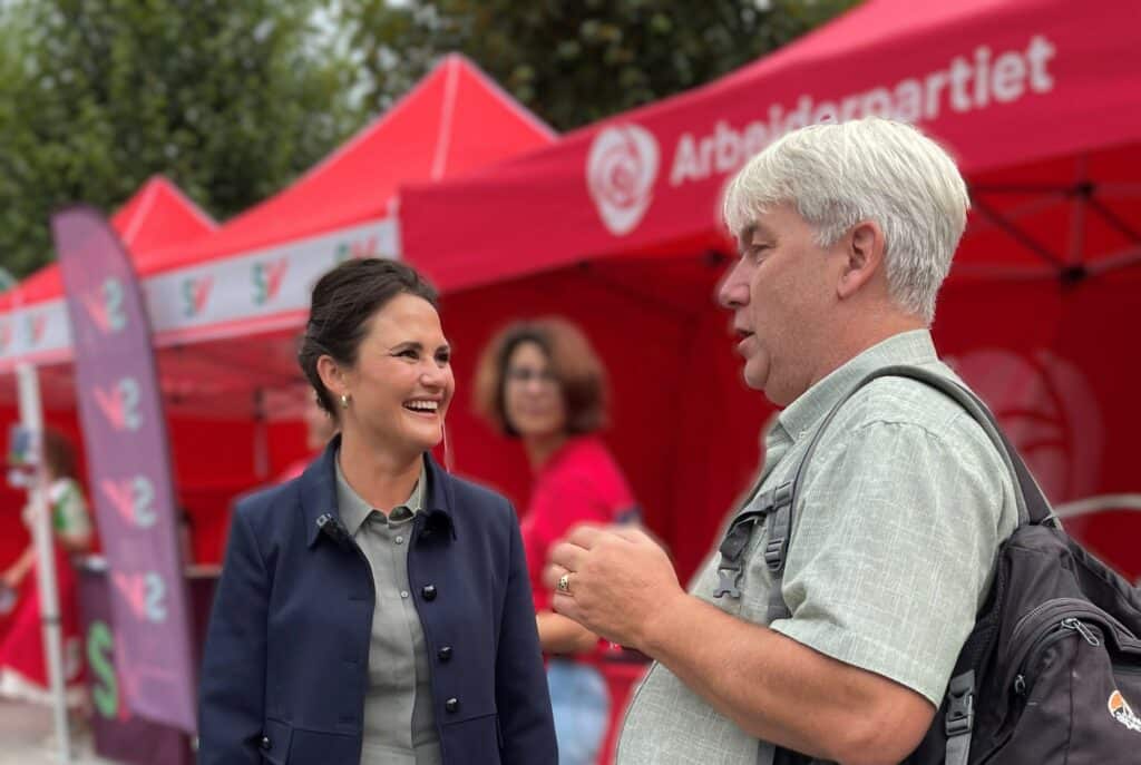En kvinne og en mann snakker og smiler i nærheten av røde Arbeiderparti-telt på et utendørsarrangement. Andre mennesker og bannere er synlige i bakgrunnen.