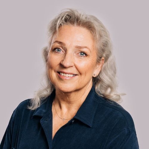 Eldre kvinne med grått hår, smilende, iført mørkeblå skjorte med krage, poserer foran en vanlig lys bakgrunn.