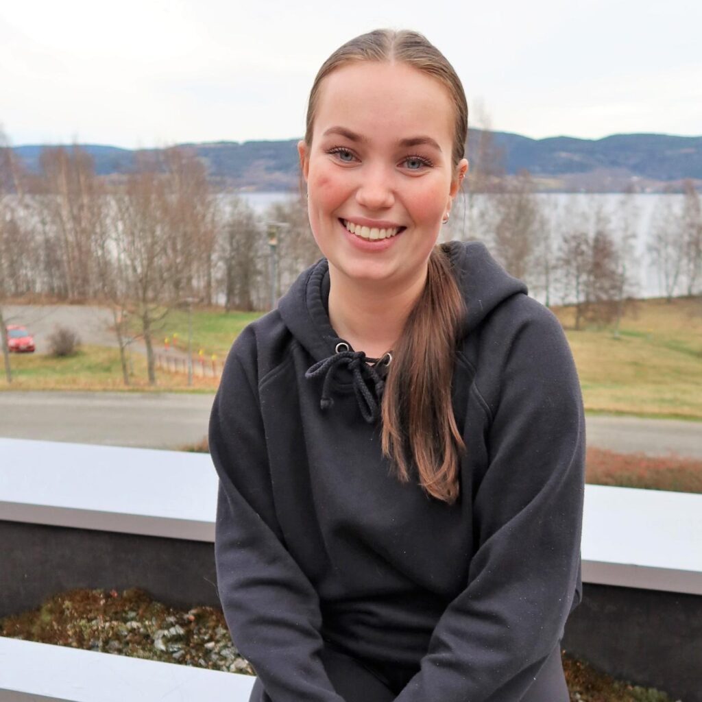 Ung kvinne med brunt, bakoverbøyd hår og svart hettegenser sitter utendørs og smiler, med en innsjø og trær i bakgrunnen.