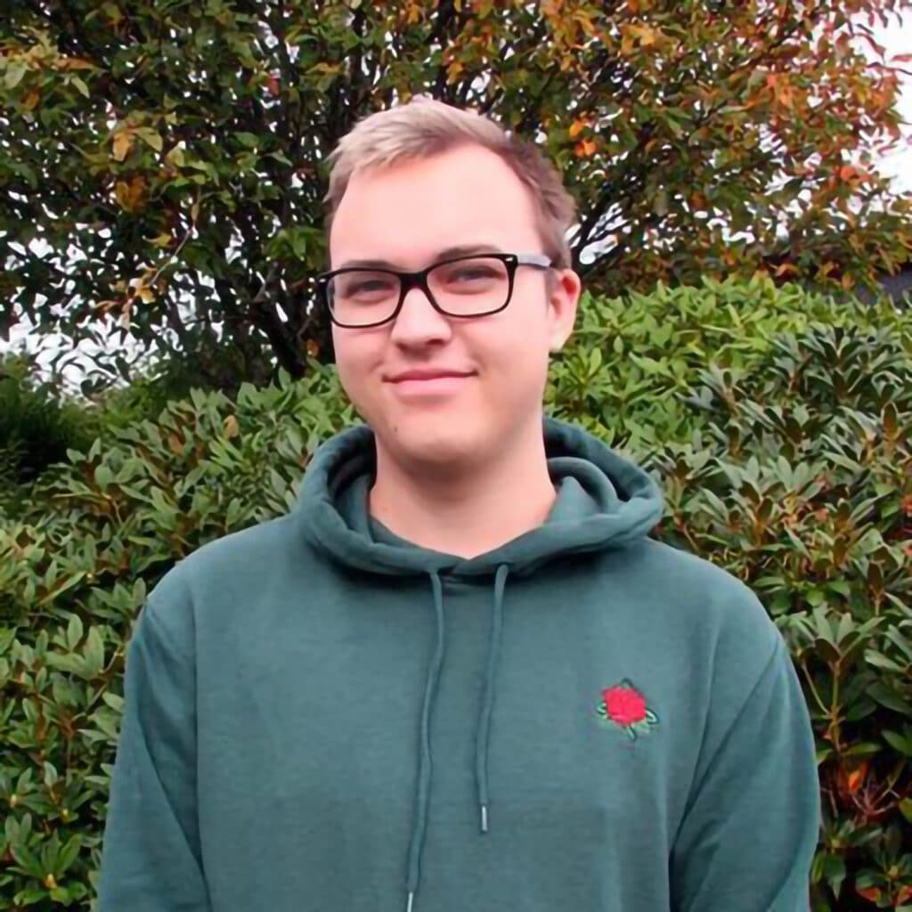 En person med briller og grønn hettegenser med et rødt roseemblem står foran tett grønt og et tre med høstløv.