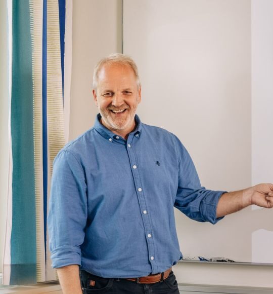 En mann i blå skjorte står smilende foran en tavle, med den ene armen utstrakt og pekende til siden.