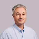 En middelaldrende mann med kort, grått hår og lyseblå skjorte, med et lett smil, mot en vanlig, lysegrå bakgrunn.