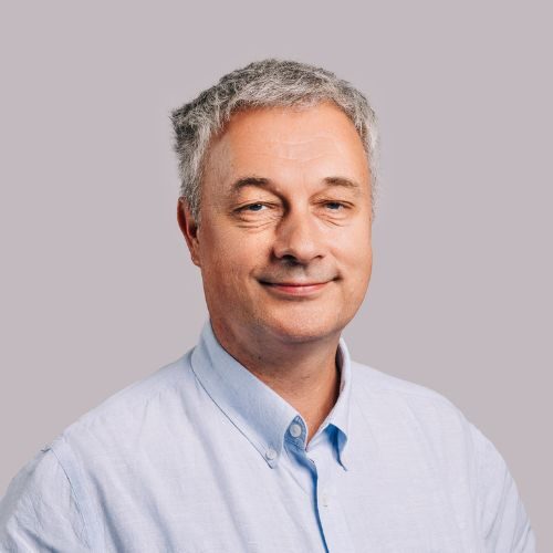 En middelaldrende mann med kort, grått hår og lyseblå skjorte, med et lett smil, mot en vanlig, lysegrå bakgrunn.